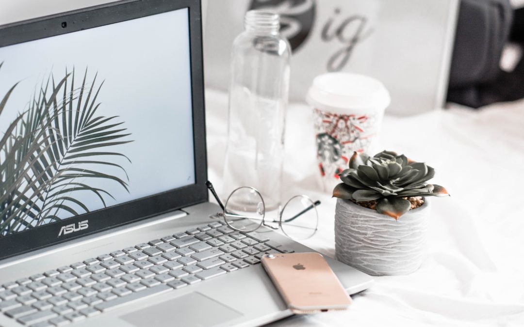 A laptop is open, and turned on. A pink iPhone is placed on the keyboard. A bottle of water, paper coffee cup, reading glasses, and a small potted succulent are to the right of the laptop.