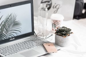 A laptop is open, and turned on. A pink iPhone is placed on the keyboard. A bottle of water, paper coffee cup, reading glasses, and a small potted succulent are to the right of the laptop.