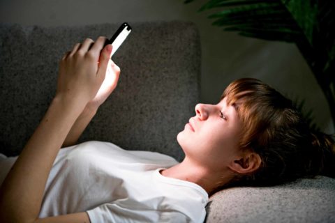 A teen girl is lying on a sofa and holding her phone. She is looking up at the screen, which is shining a white light on her face.