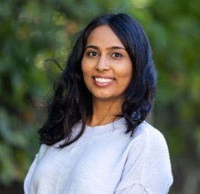 Dr. Aneela Cox smiles, facing the camera, with trees blurred in the background.