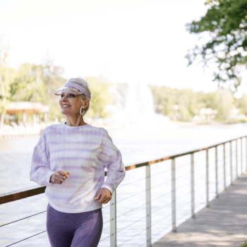 A woman is smiling and walking next to a railing. She is wearing a clear visor cap, and has grey hair. The background is blurry.