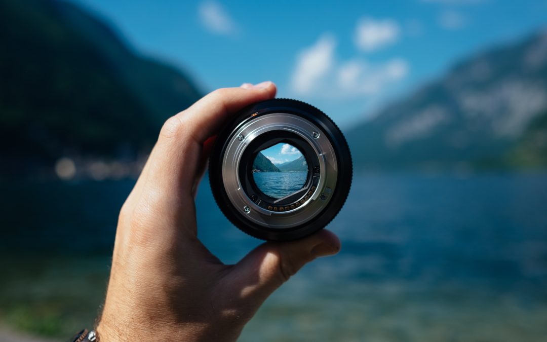 A hand holds up a camera lens focused on a lake and mountains in the distance. The background is blurry.