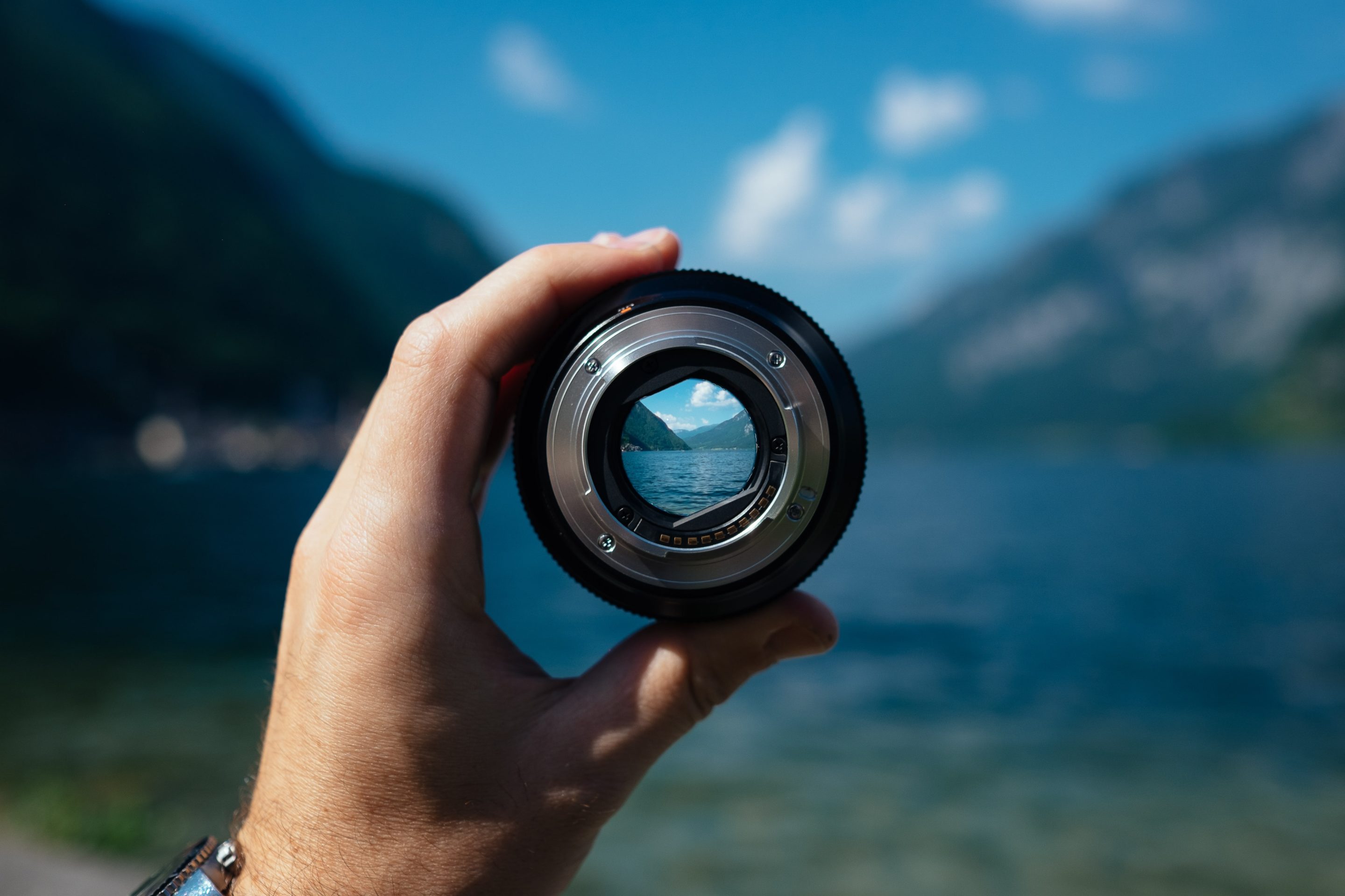 A hand holds up a camera lens focused on a lake and mountains in the distance. The background is blurry.