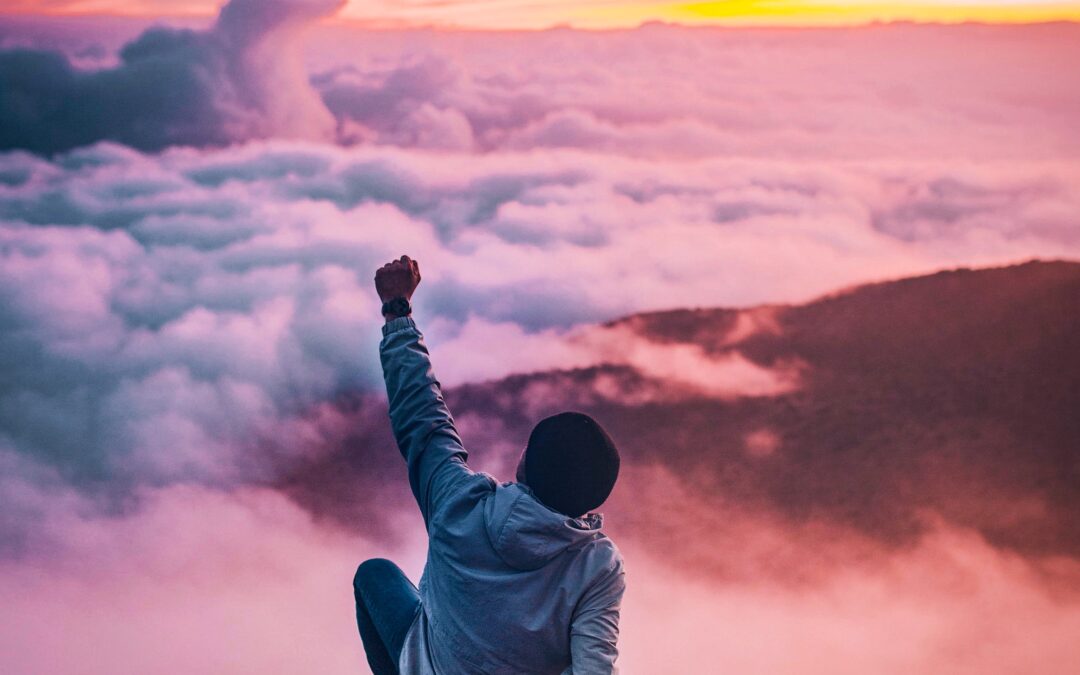 A person sits on top of a mountain peak with a fist raised to the sky. The horizon of mountains is obscured by clouds and the sky is full of bright colors.