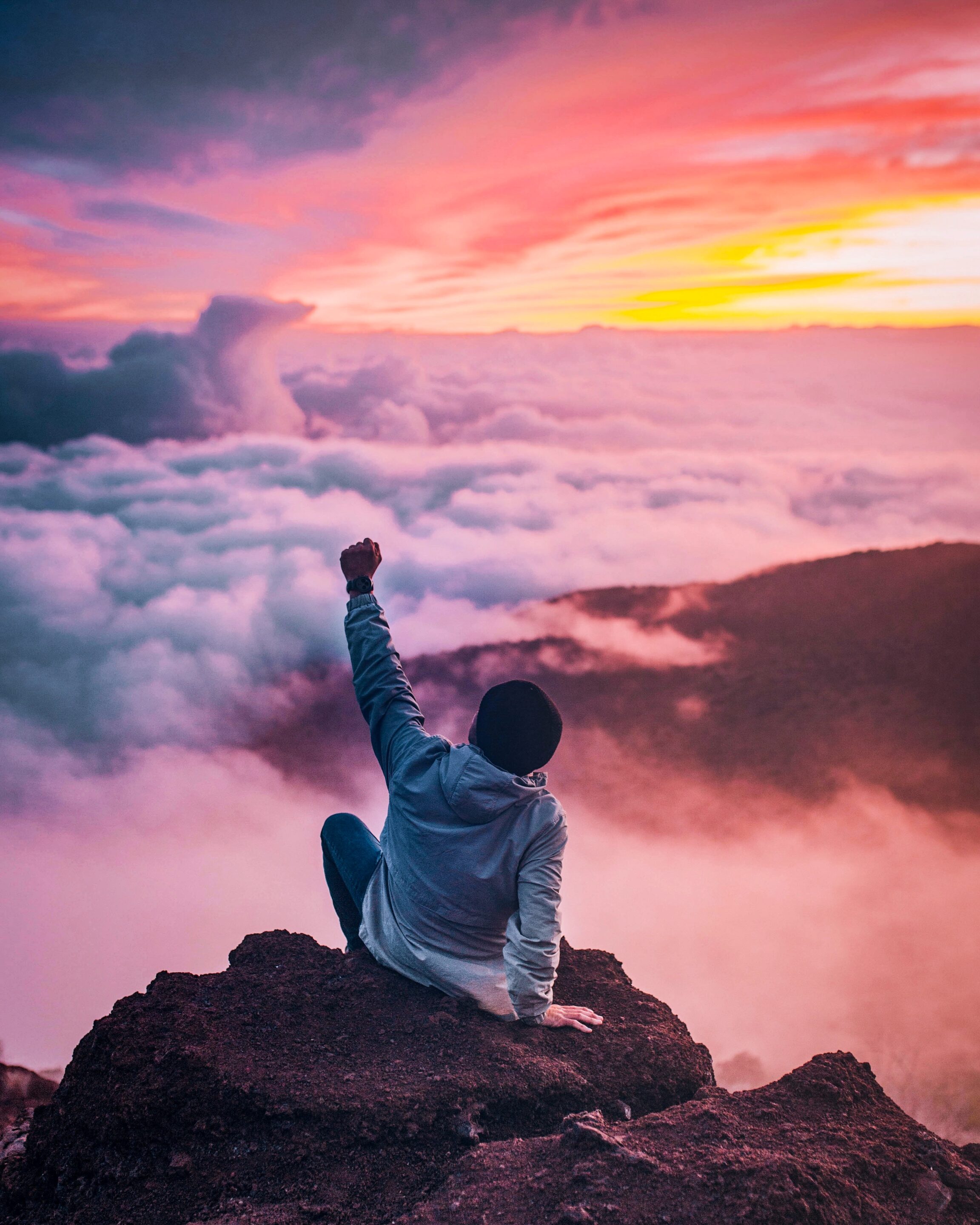 A person sits on top of a mountain peak with a fist raised to the sky. The horizon of mountains is obscured by clouds and the sky is full of bright colors.