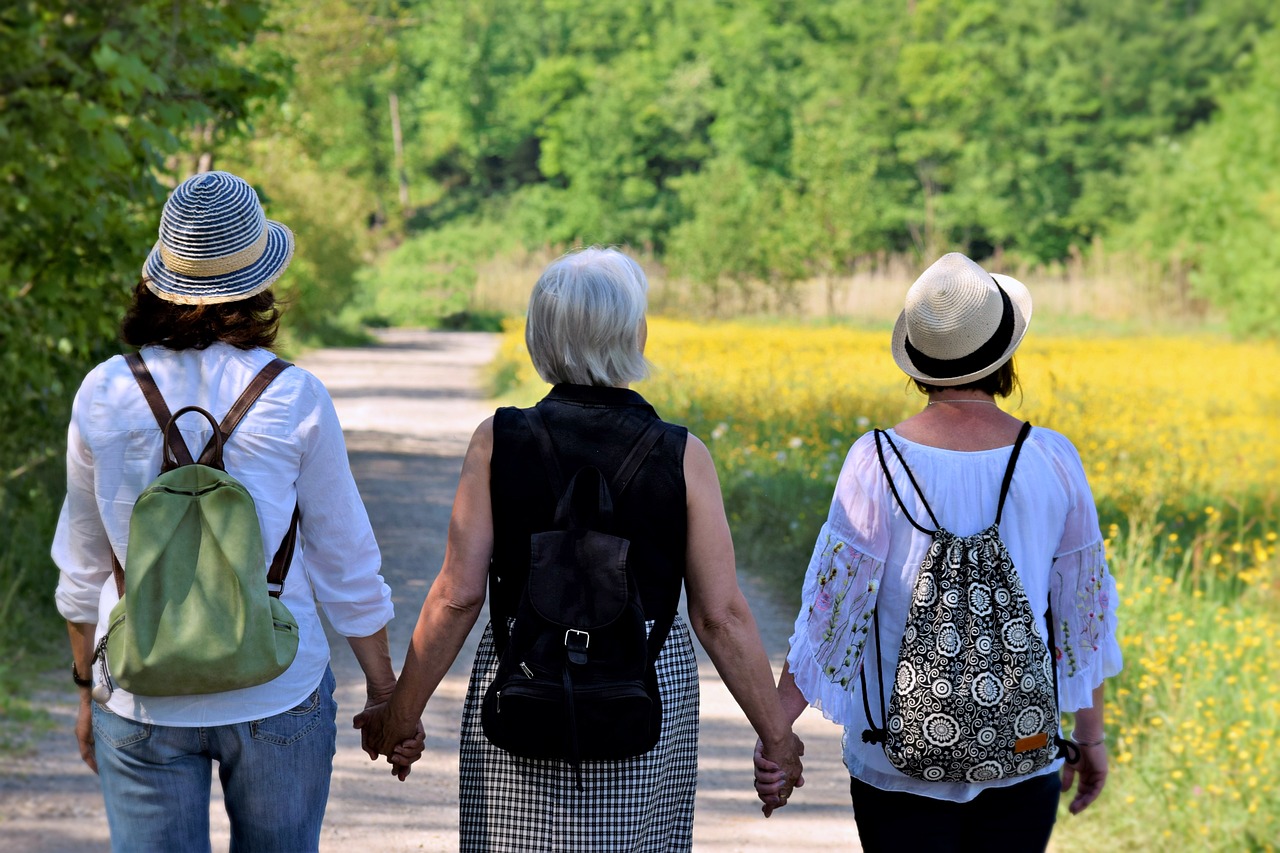 Three women of varying ages walk with their backs to the camera, holding hands, on a trail with sunlit fields and trees in the distance.