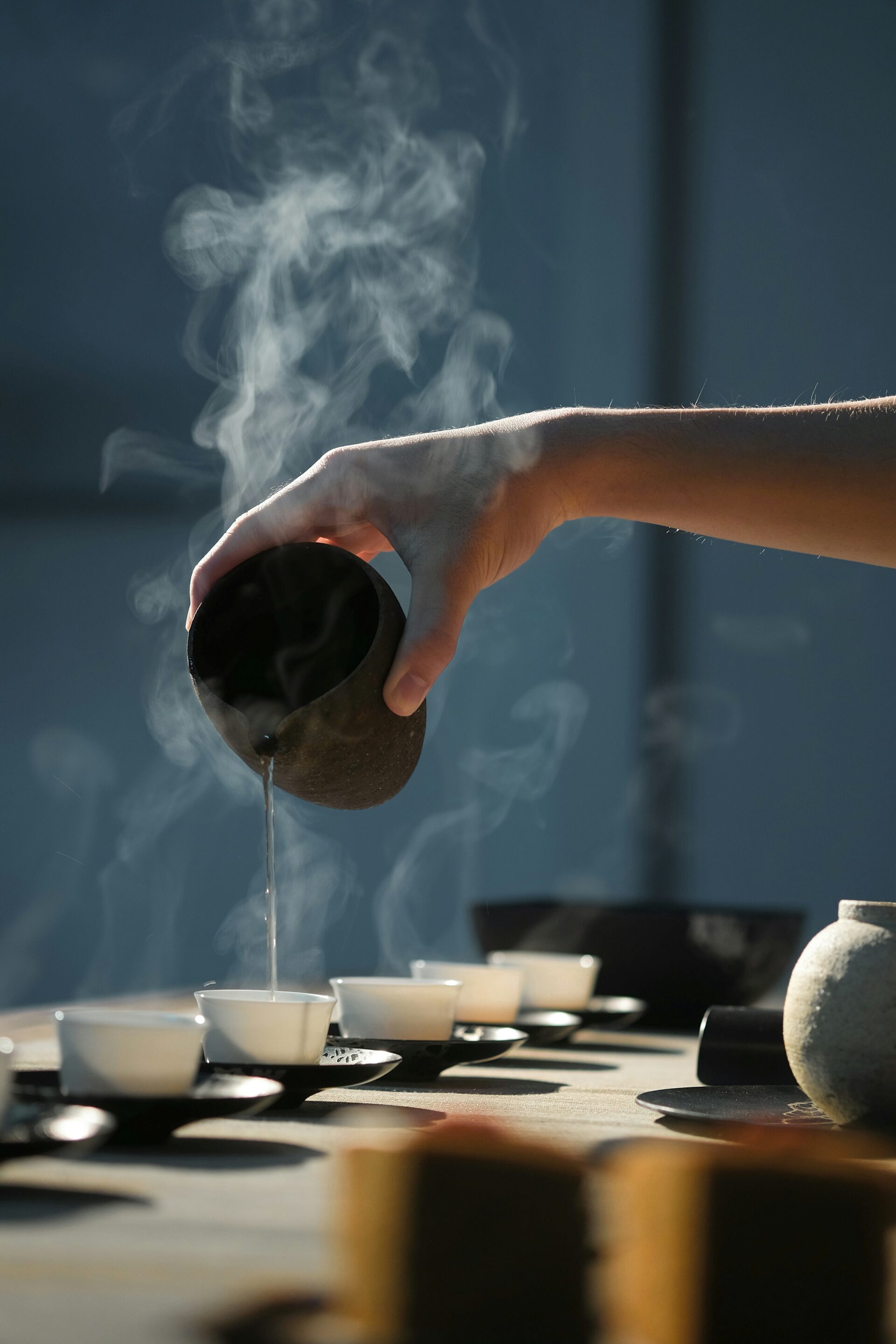 In a tea ceremony, a person's arm and hand are pouring hot tea into a row of small ceremonial tea cups.