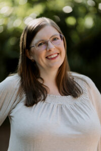 A woman wearing glasses smiles. The background is out of focus, but appears to be sunlit trees.