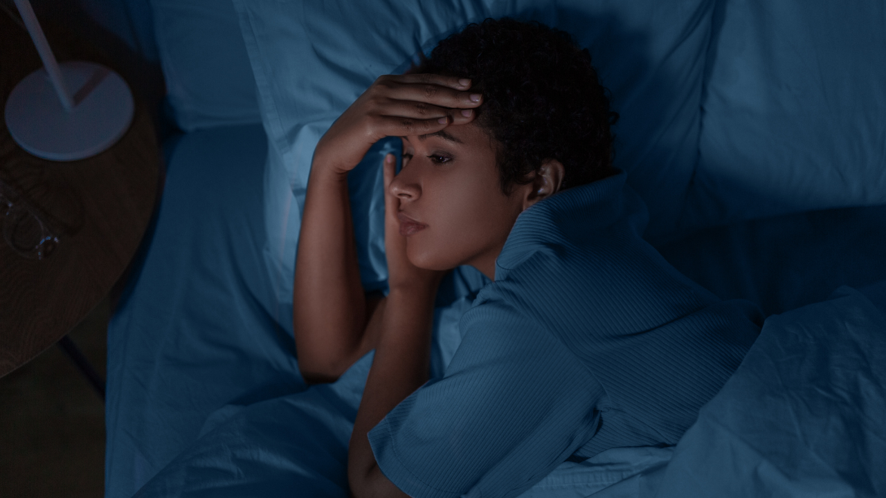 A woman lies in bed, trying to sleep. It is dark, with her head on a pillow, a hand on her forehead, and her eyes open.