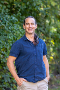 A man stands in front of greenery wearing a blue shirt. He is smiling.