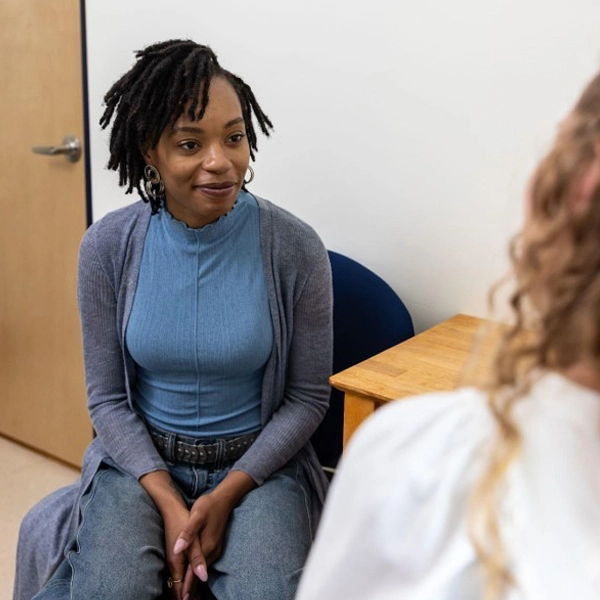 A woman with short dreadlocks, wearing a blue top and gray cardigan, sits on a chair in an office, attentively listening to another person whose back is to the camera.